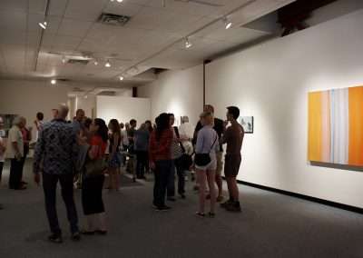 People viewing and discussing art in a brightly lit gallery with white walls and modern paintings. various groups are engaged in conversations across the room.