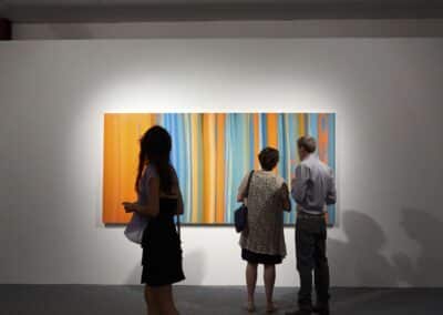 Three people viewing a large, horizontal abstract painting with bold orange and blue stripes in an art gallery with white walls.