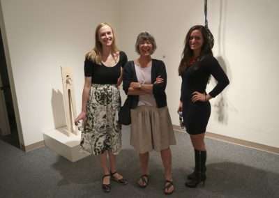 Three women smiling and posing together at an art gallery. one in a floral skirt and sandals, one in a beige skirt and sandals, and one in a black dress and high boots.