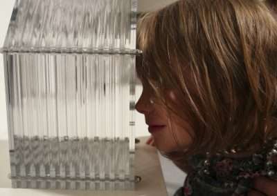 A young girl with light brown hair, wearing a floral blouse, closely examines a transparent, geometric sculpture displayed on a white pedestal in a gallery setting.