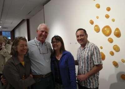 Four people smiling at an art gallery, standing in front of a wall decorated with round, amber-colored art pieces.