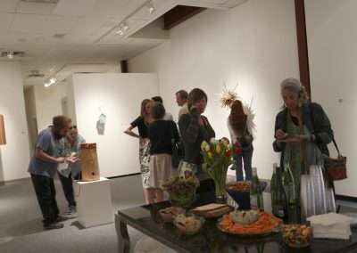 People viewing artwork in a gallery with displays of sculptures and a table with snacks and beverages in the foreground.