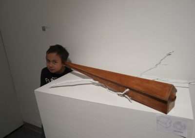 A young child peeks from behind a wooden art installation on a white pedestal in a minimalist gallery, expressing curiosity.