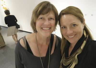 Two women smiling at the camera in an art gallery, one with short hair and a black top, the other with long hair and a black dress. other visitors are observed in the background.