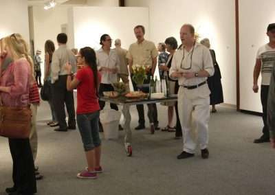People mingling in an art gallery, holding drinks and conversing. a woman in the foreground carries a tray of hors d'oeuvres. artworks are displayed on the white walls around the room.