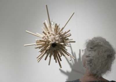 A woman with gray hair examines a spherical, wooden sculpture hanging from the ceiling, composed of numerous sticks protruding in all directions.
