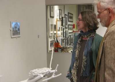 A mature couple examines a small, intricate white sculpture displayed on a pedestal in an art gallery, with framed artwork hanging on walls around them.