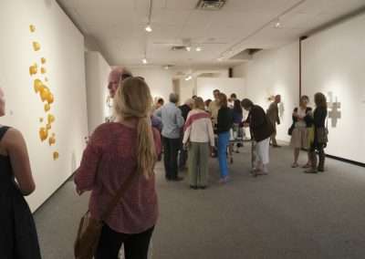 Visitors observing diverse artworks at an art gallery exhibition, featuring wall-mounted pieces and a lively crowd engaged in conversation.