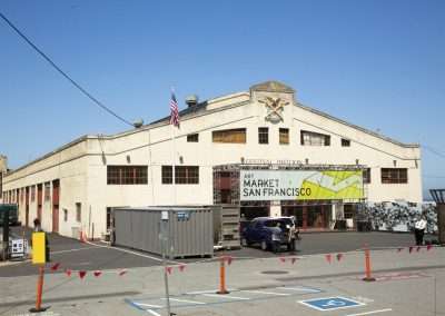 Exterior view of a large, aged industrial building adorned with banners for the art market san francisco, under a clear blue sky. a car is parked outside, and safety cones line the foreground.