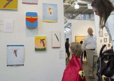 A child in a superhero cape runs excitedly through an art gallery with abstract paintings on the wall, while an adult, likely a parent, looks on from beside a stroller.