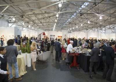 A bustling indoor art fair with people viewing various artworks in a spacious, brightly lit hall with white walls. visitors mingle and converse, some standing by artworks and others near food stalls.