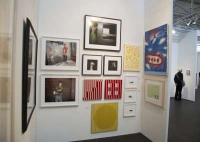 An art gallery display featuring a variety of framed artworks, including photographs and abstract pieces, on white walls, with a few visitors observing the art.