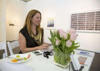 A smiling woman sitting at a desk in a bright office, with a vase of pink tulips, colorful brochures, and a digital tablet in front of her.