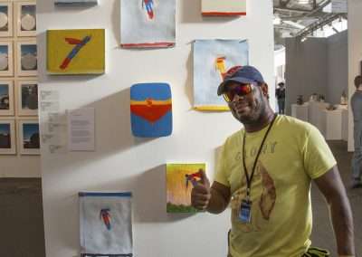 A man in a yellow t-shirt and a cap stands smiling next to a display of colorful abstract paintings on a white gallery wall, giving a thumbs up.