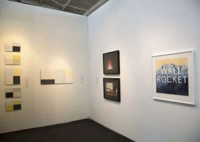 An art gallery wall featuring a diverse array of paintings and photographs, including modern abstract pieces and a framed photograph of a mountain labeled "rocket.