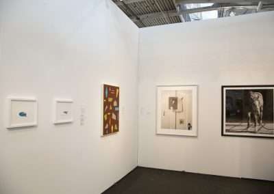 A bright, modern art gallery with white walls displaying five diverse artworks, including paintings and a photograph, in a clean, well-lit exhibition space.