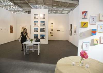 A woman standing at a desk in a bright art gallery featuring diverse paintings and photographs displayed on white walls.