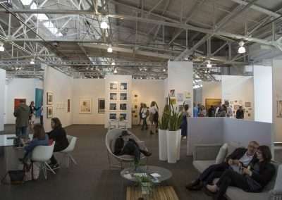 Spacious art fair with visitors exploring various gallery booths displaying paintings, photographs, and art installations inside an industrial-style venue with high ceilings.