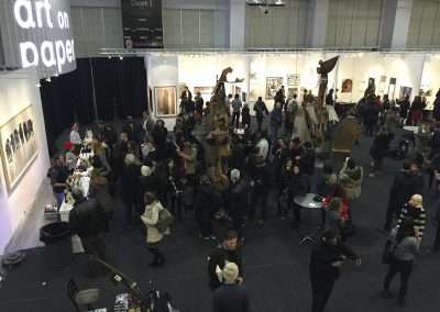 An indoor art fair scene with many visitors viewing a series of displays and art works. the event is bustling with activity in a large, well-lit hall.