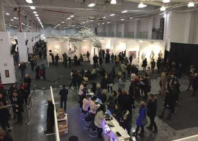 Overhead view of a bustling contemporary art fair with visitors viewing various artwork displays in a large, well-lit exhibition hall.