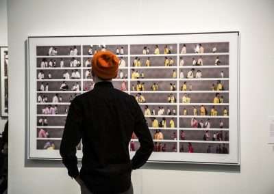 A person in a black jacket and orange beanie views a large framed artwork featuring multiple rows of photographic portraits, each depicting various individuals in different colored outfits.