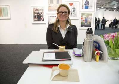 A woman wearing glasses and a smile sits at a small table with a cup of soup, a notebook, and a laptop in an art exhibition space. tulips and a thermos are also on the table.
