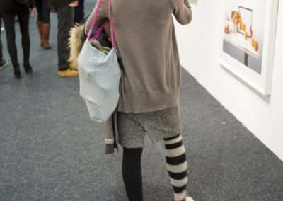 A woman in layered clothing and striped socks views art at a gallery, her back to the camera, with a tote bag and a camera slung over her shoulder.