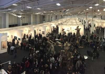 Panoramic view of a busy indoor art exhibition with numerous attendees viewing various artworks displayed in well-lit white booths.