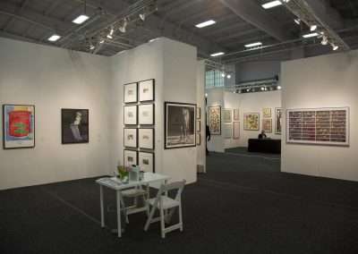 An art gallery interior with white walls displaying various framed artworks, including photographs and paintings. a small seating area with a white table and chairs is situated in the foreground.