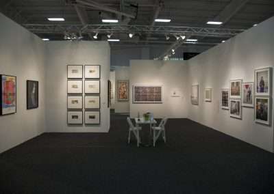 Modern art gallery exhibition space with various framed artworks on white walls, and a small seating area with a table and two chairs in the center.