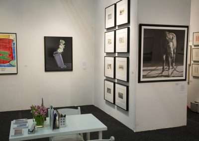 An art gallery interior with various paintings and photographs displayed on white walls, including a large horse photo, and a small seating area with a desk and flowers.