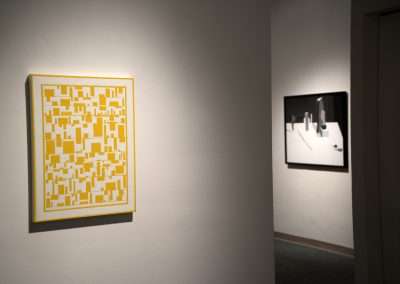 A yellow and white abstract painting is displayed on a gallery wall, with another artwork partially visible in the background, creating a focused and tranquil atmosphere.