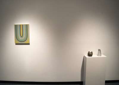 A modern art gallery with a brightly colored striped painting on a white wall and two sculptures on pedestals in a dimly lit room.