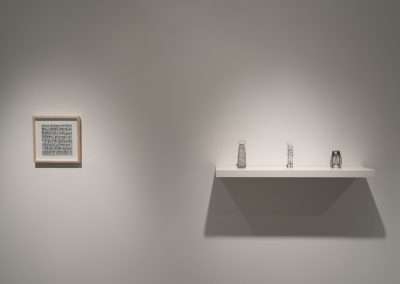 A minimalist art gallery display featuring a framed textual artwork on the left wall and a shelf with three small sculptures on the right, all under soft lighting.