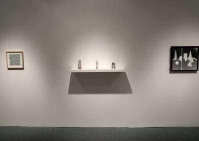 A minimalist art exhibition display featuring one small textual artwork on the left wall, a shelf with three small sculptures in the center, and a graphic black and white painting on the right wall.