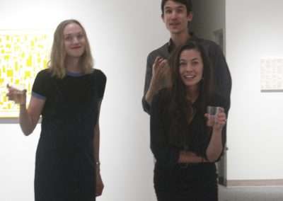 Three young adults at an art gallery, one man standing behind two women, all smiling. artwork on the walls and a white, neutral setting.