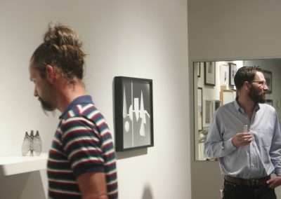 Two men observing various artworks in a gallery, with one holding a drink. the artwork includes framed abstract and architectural designs. the setting is well-lit and modern.