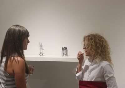Two women, one with straight dark hair and the other with curly blond hair, converse in an art gallery, standing beside a small sculpture exhibit.