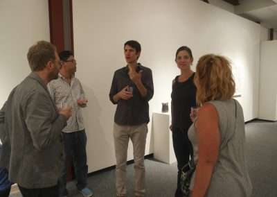 Five adults standing and conversing in an art gallery, holding drinks, with artwork displayed in the background.