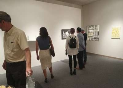 People viewing art in a gallery, with some standing close to the walls examining pictures, and one person walking to the left foreground.