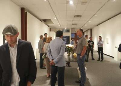 Visitors engaging at an art gallery exhibition, with some individuals closely observing artworks and others in conversation. the gallery is lit with soft overhead lighting.