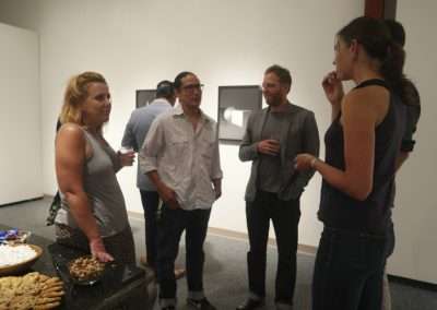 Five adults chat and enjoy refreshments at an indoor gallery event, with a table of snacks in the foreground.