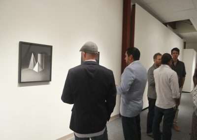 People viewing a modern abstract artwork in a white-walled gallery corridor. one man, wearing a hat, is closely observing the piece.