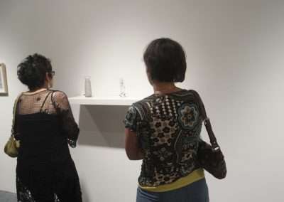 Two women viewing artwork in a gallery, standing before a shelf displaying small sculptures. the atmosphere is calm and contemplative.