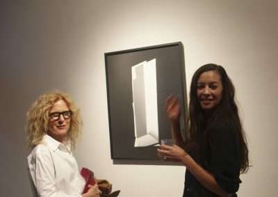 Two women smiling and standing next to a framed abstract geometric artwork in a gallery. one woman appears to be explaining or gesturing towards the artwork.