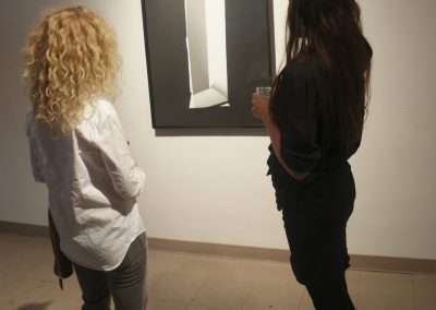 Two women, one with curly blonde hair and the other with long brunette hair, standing in an art gallery observing a framed abstract monochrome artwork.