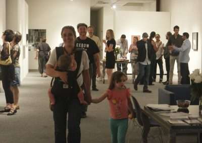 A bustling art gallery scene where diverse groups of people, including a woman carrying a baby and holding hands with a young girl, engage in viewing artwork and social interactions.