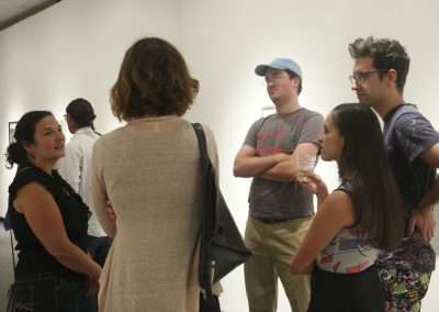 A group of five adults engaging in conversation in an art gallery, with artworks hanging on the white walls in the background. two men and three women, casually dressed, appear relaxed and interested.