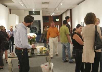 People mingling at an indoor gallery event, with a man examining a flower arrangement on a table in the foreground, and other attendees conversing in the background.