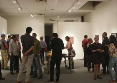 People mingling and viewing artworks at an indoor art gallery exhibition. the room is well-lit and features white walls displaying various framed pieces.